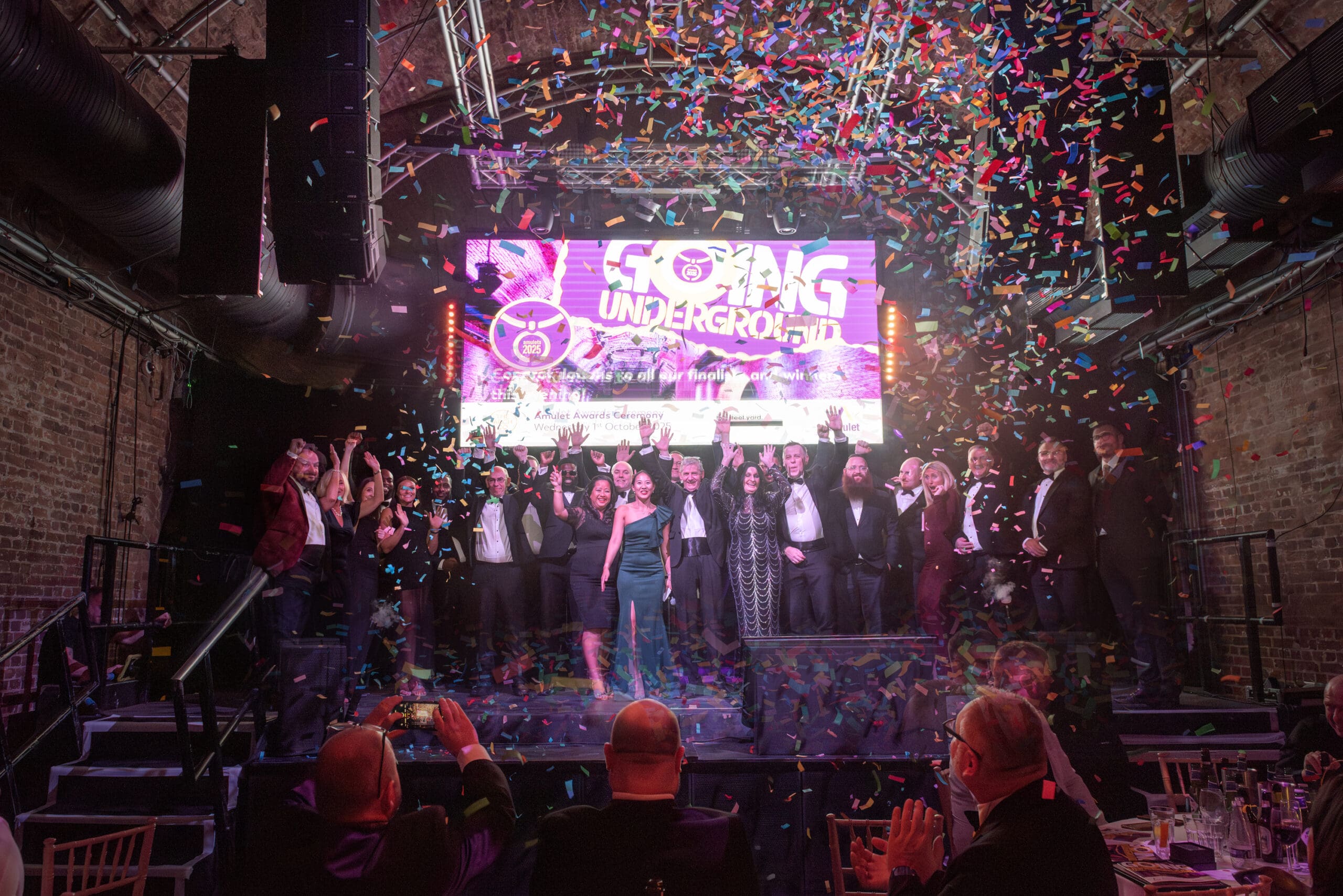 Group of people standing on stage, cheering, whilst confetti falls